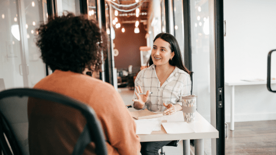 Two female co-workers sat across a desk discussing employee wellbeing
