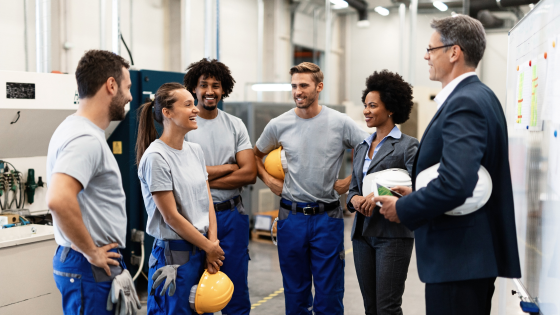 A diverse group of colleagues in workwear chatting together in a factory setting, with a manager in a suit joining the conversation